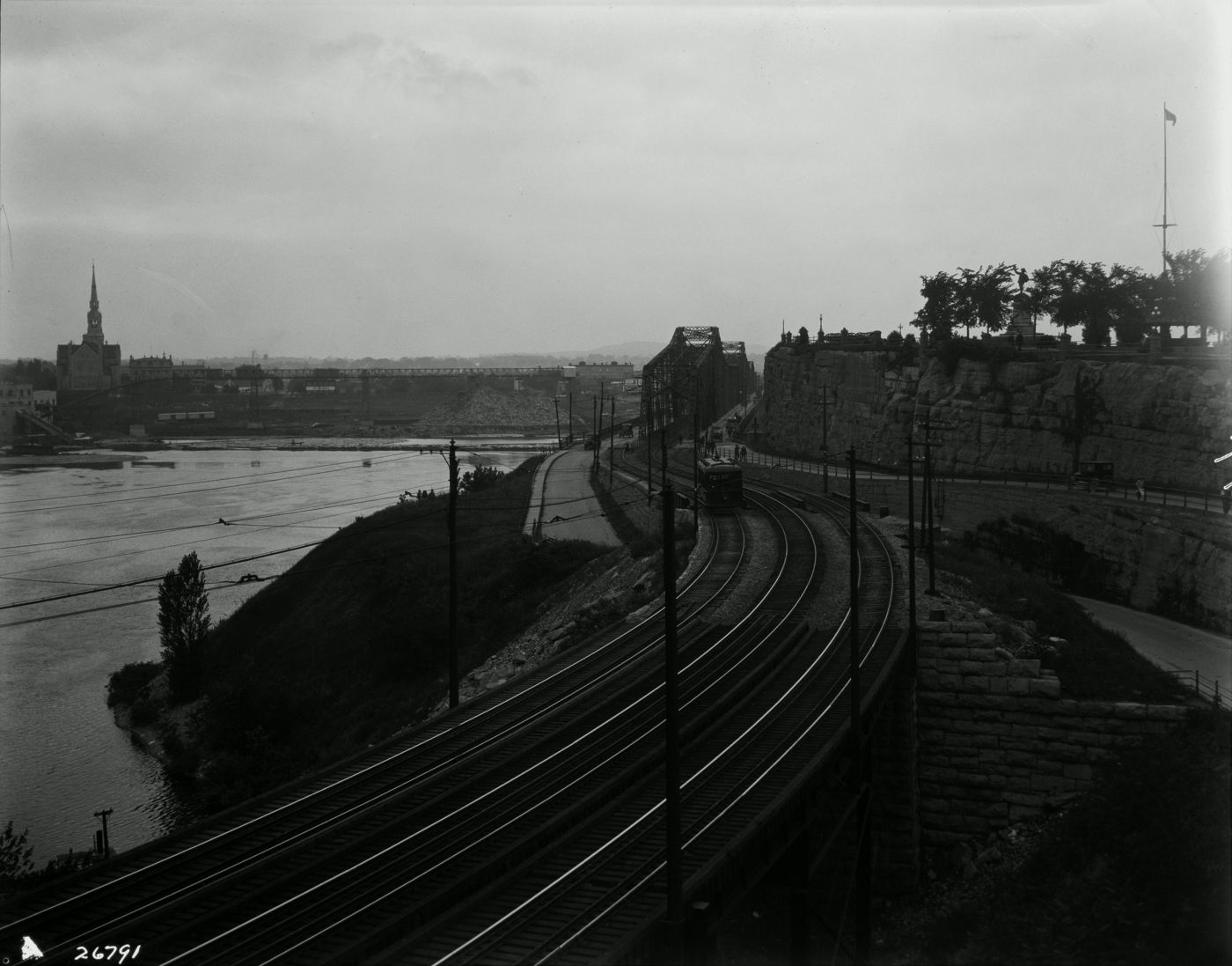 Black and white photograph of a riverside landscape. In the foreground, three railways lead to an iron bridge. On the other side of the river, an industrial landscape dominated by an imposing church.