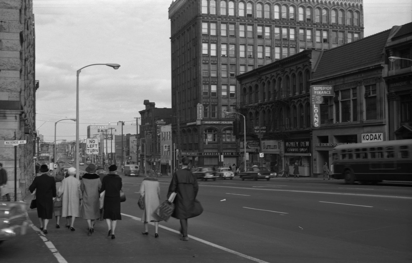 Black and white photograph of a busy street with multi-storey buildings and billboards. In the foreground, a group of well-dressed pedestrians crosses a side street.