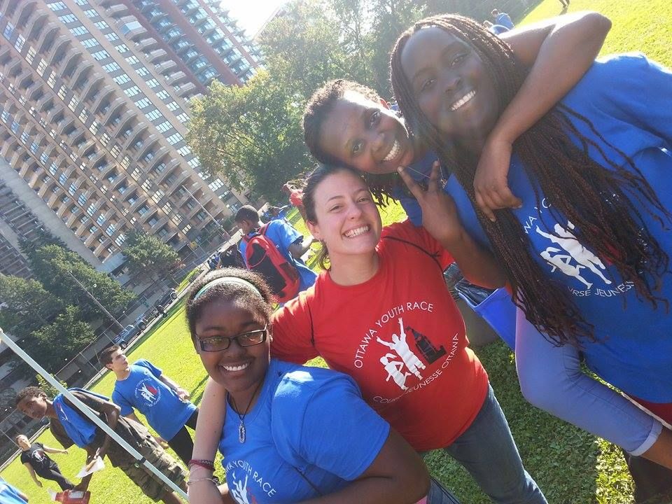 Photographie oblique en couleur de trois adolescentes noires en tee-shirt bleu et d’une jeune adulte blanche en tee-shirt rouge sur lequel apparaissent le logo et le nom de l'événement Ottawa Youth Race/Course Jeunesse Ottawa. À l’arrière-plan, un champ clôturé occupé par d’autres jeunes et de grands immeubles résidentiels.