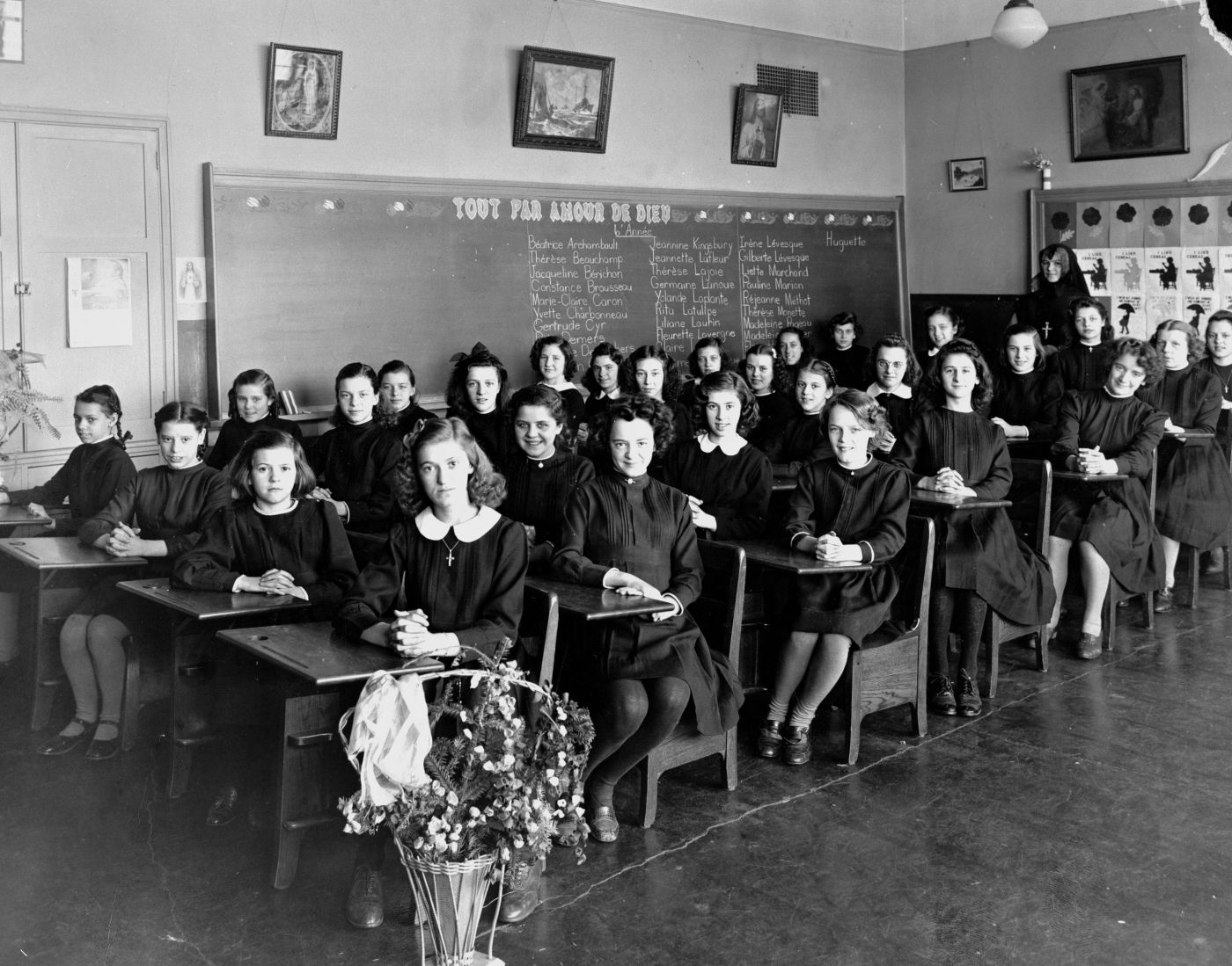 Photographie en noir et blanc d’une salle de classe vue en angle. Une trentaine de jeunes filles en robes noires sont assises à des pupitres. La classe compte sept rangées de quatre pupitres. Une religieuse est debout au fond de la salle de classe.