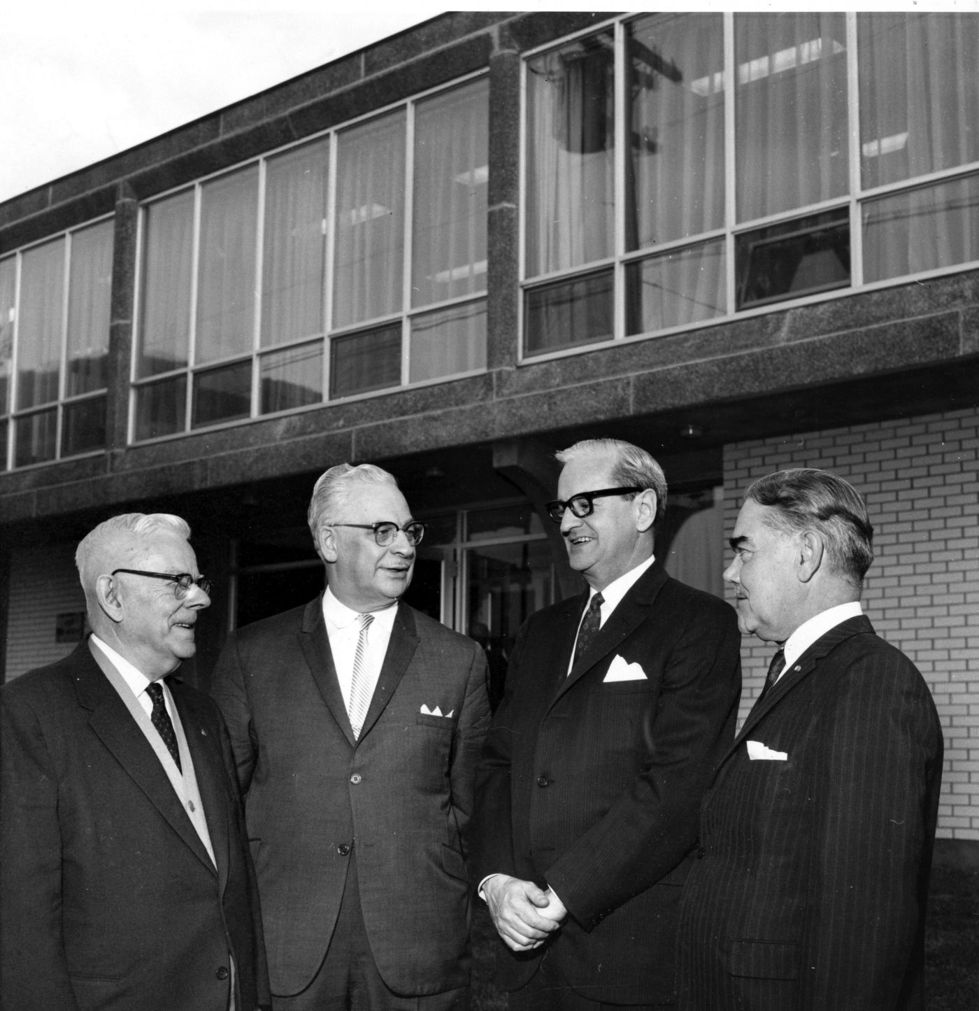 Photographie en noir et blanc de quatre hommes d’un certain âge portant costume et cravate, qui discutent devant un édifice à deux étages en brique. Le deuxième étage dispose de belles fenêtres.