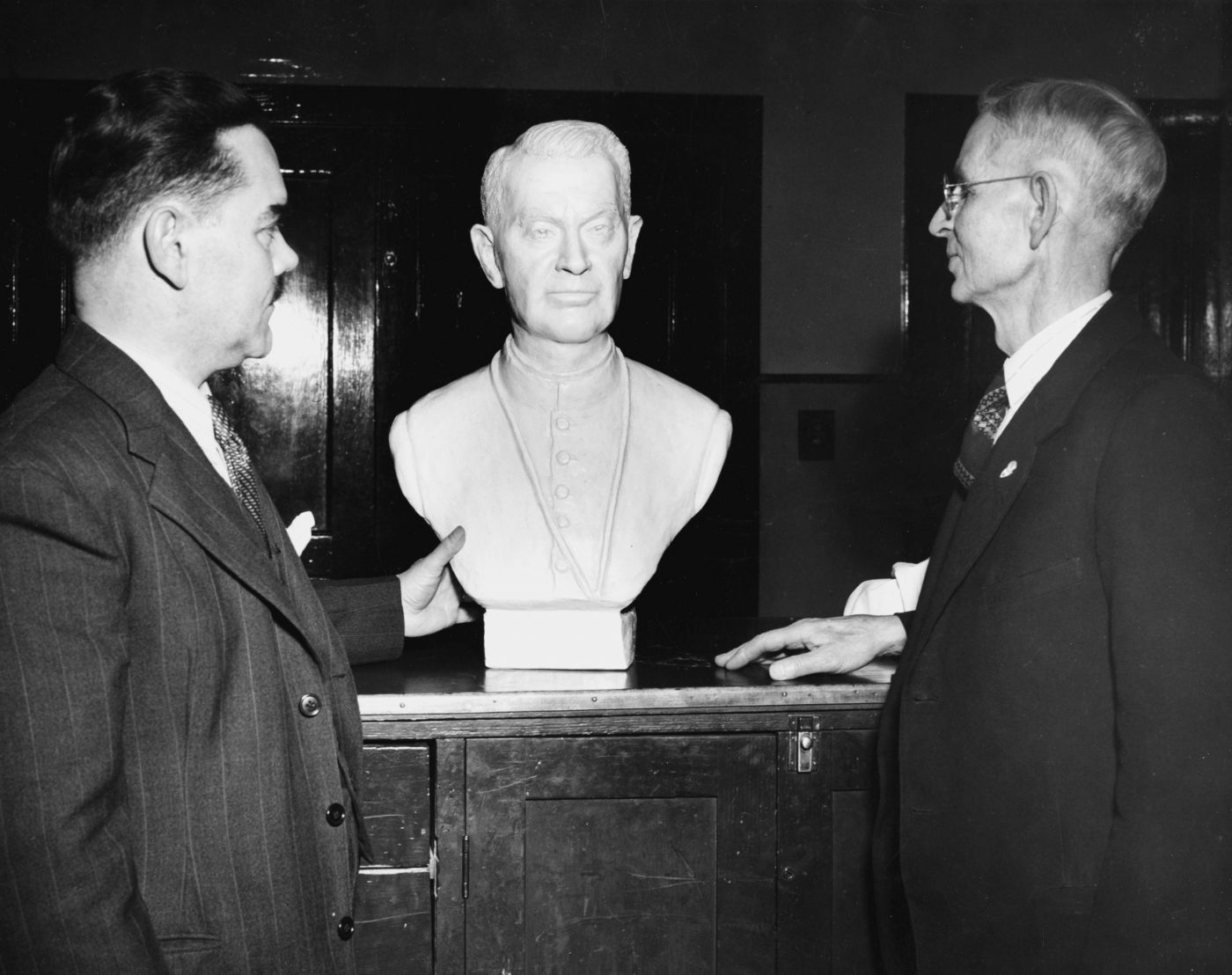 Photographie en noir et blanc de deux hommes, en costumes et cravates, debout de chaque côté d’un buste en plâtre d’un religieux, d’âge mûr. Le premier, d’âge moyen et moustachu, touche le bas du buste de sa main gauche; le second, d’âge mûr et portant lunettes, pose sa main droite sur le coffre où est posé le buste.