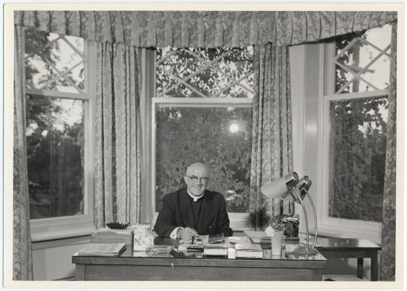 Photographie en noir et blanc d’un religieux assis à un bureau, un stylo à la main. Il porte des lunettes et son crâne est dégarni. II a relevé la tête et sourit à la caméra. Derrière lui, trois grandes fenêtres ornées de rideaux fleuris, qui laissent voir de grands arbres.