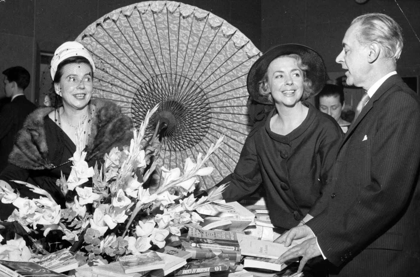 Black and white photograph of an older man, and two elegantly dressed, smiling middle-aged women. They are standing around a table with books piled around a large bouquet of flowers and an open parasol.
