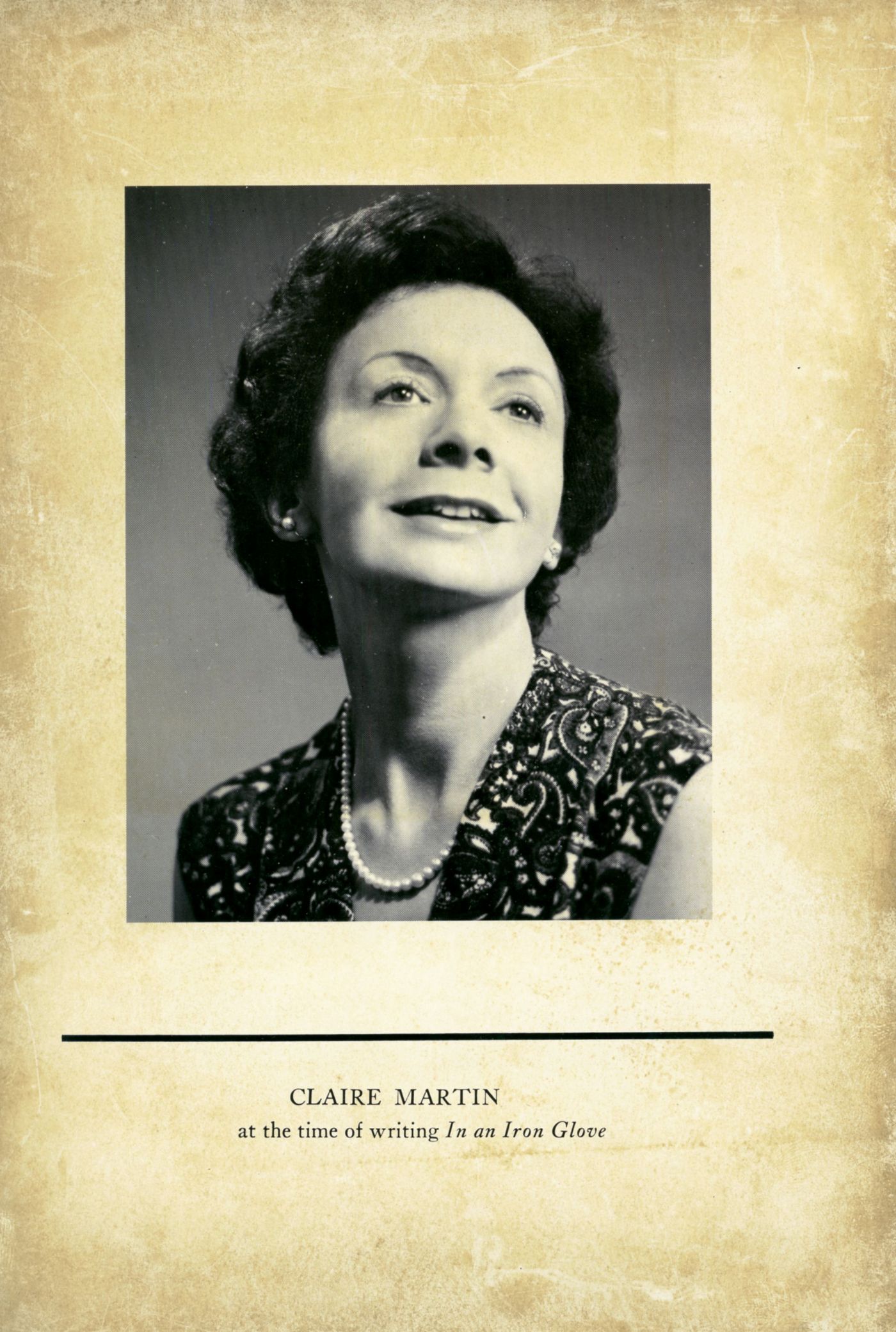 Black and white studio photograph of a middle-aged woman, smiling, face turned slightly to the left. She has short, slightly wavy, dark brown hair, and she wears a sleeveless, patterned garment and pearl necklace. An English inscription appears below the image.