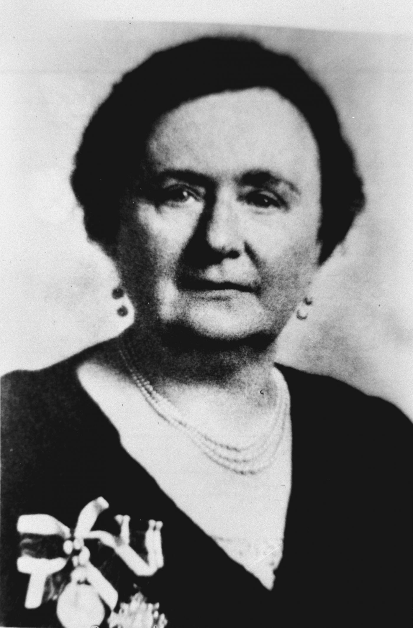 Photographie en noir et blanc, gros plan, prise en studio d’une femme d’âge mûr, vue de face. Elle porte des boucles d’oreille et un collier de perles enfilées sur trois rangs. Elle porte aussi des fleurs à la boutonnière. Elle affiche un air sérieux.