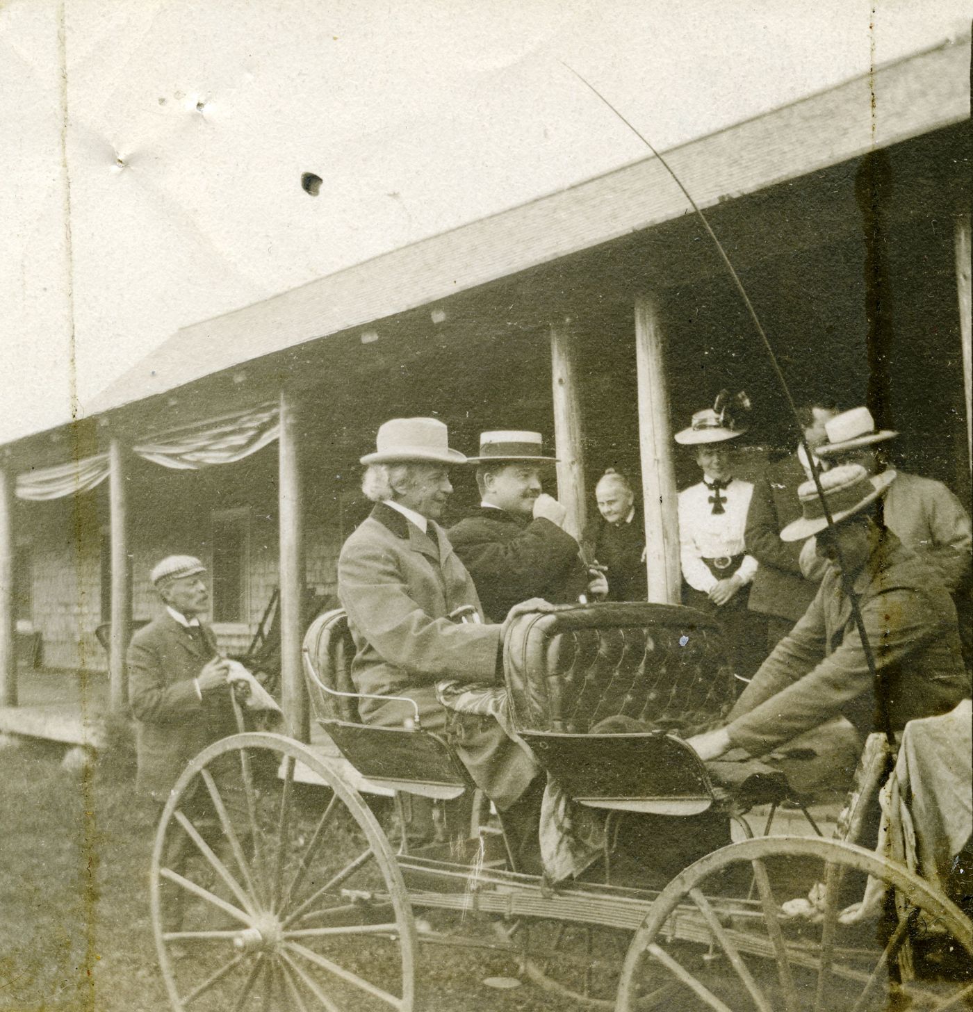Photographie en noir et blanc de deux hommes bien mis, assis dans un boghei devant un bâtiment en bois. Les cheveux blancs du premier sont visibles malgré son chapeau. Le second, plus jeune, porte la moustache.  Un troisième homme prépare le boghei. Quelques personnes les observent.