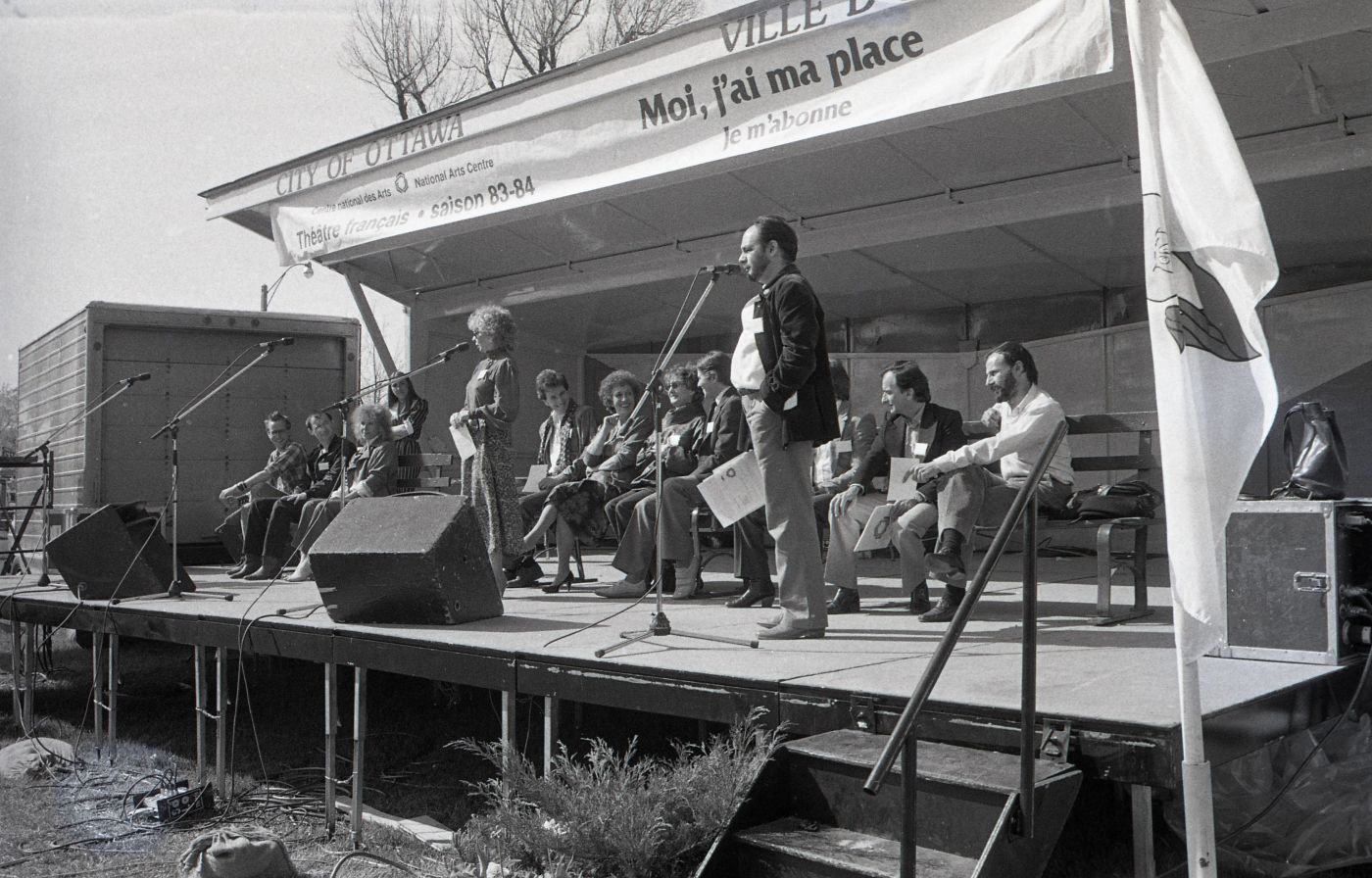 Black and white photograph of a middle-aged man and woman standing at microphones on an outdoor stage. Behind them, a dozen people sitting on benches. On a banner: “Centre national des Arts–National Arts Centre; Théâtre français saison 83-84; Moi, j’ai ma place; Je m’abonne.”