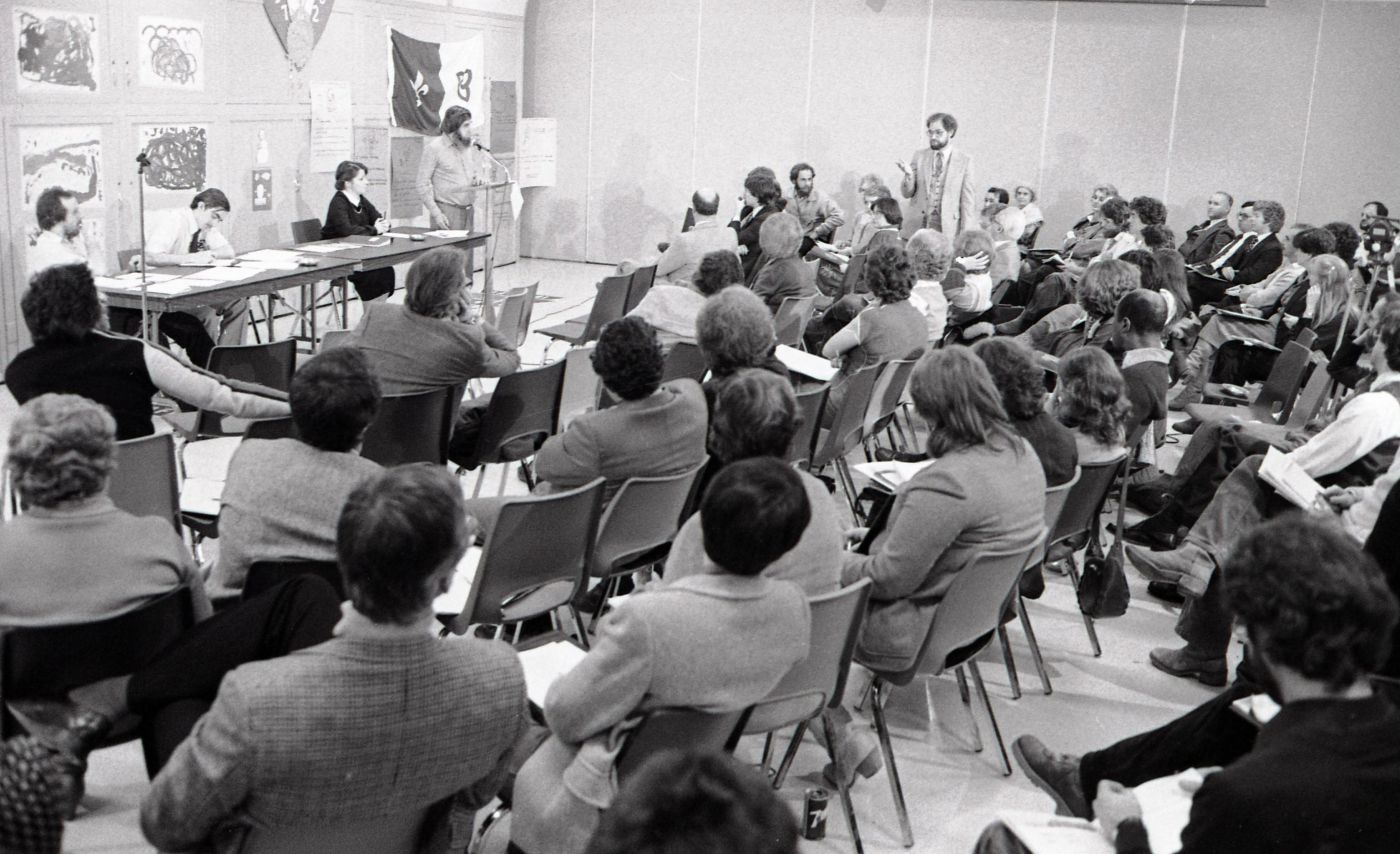 Photographie en noir et blanc d’une réunion à laquelle participent des dizaines de personnes, vues de dos. Devant elles, trois intervenants, dont une femme, sont assis à une table sur laquelle sont déposés des documents. Un quatrième est debout au micro. Il s’adresse visiblement à un des participants, qui s’est levé durant l’échange. Le mur qui fait face à la salle est tapissé d’affiches. Le drapeau franco-ontarien y est accroché.