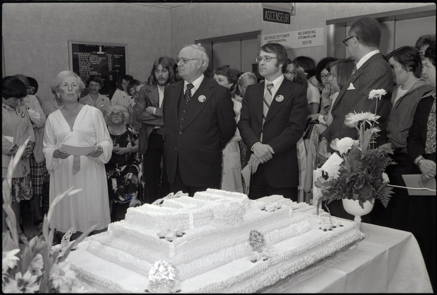 Photographie en noir et blanc d’un groupe de personnes autour d'un immense gâteau décoré du logo de l'hôpital Montfort.  Deux gerbes de fleurs ont été déposées sur la table. À l'avant-plan, trois hommes en costume arborant le même macaron et une femme vêtue avec élégance. Celle-ci tient une liasse de feuilles.