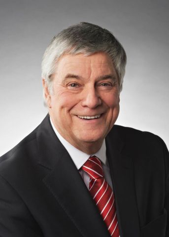 Colour photograph of an older man with greying hair, wearing a dark suit, white shirt, and striped red tie. He smiles at the camera.