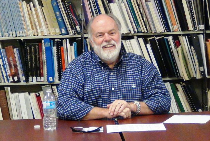 Photographie en couleur d’un homme d’âge mûr, portant la barbe. Vêtu d'une chemise carreautée, il sourit à la caméra. Il est assis à une table. Derrière lui, une bibliothèque.