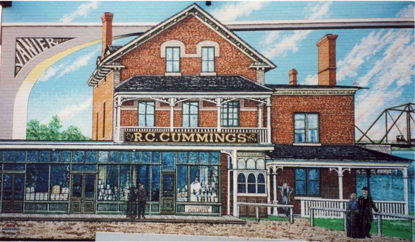 Color photograph of a mural depicting near and far views of the “R.C. Cummings” commercial building in red brick. The building, located on an island in the center of a river, is reached by a metal bridge.