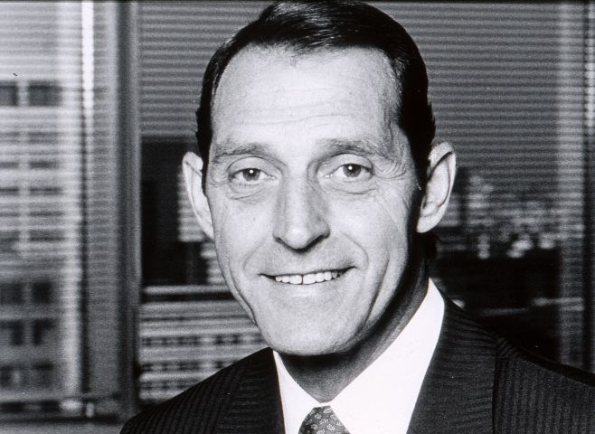 Photographie en noir et blanc d’un homme d’âge moyen souriant, en costume-cravate. L’homme est devant une fenêtre, avec vue sur des édifices en hauteur.