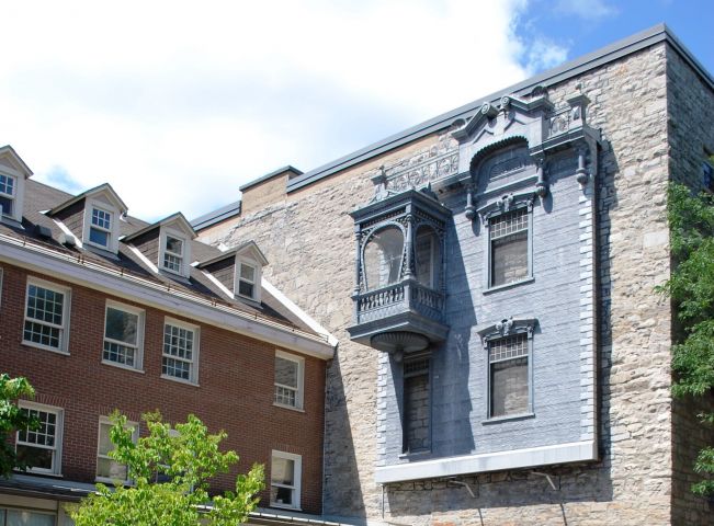 Colour photograph of an inner courtyard with a fountain in the foreground, and two buildings – one in wood, the other in stone – behind it. The metal facade of a house is fastened to the stone building, near the top of the wall.