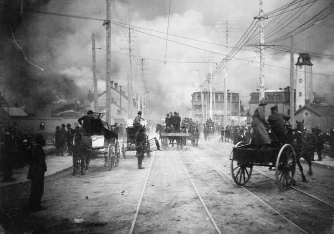 Photographie en noir et blanc d’une grande foule dans une rue menacée par la fumée. Certains sont en boghei avec leurs biens. Des pompiers arrivent en boghei.