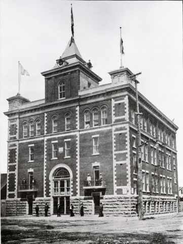 Black and white photograph of a four-storey brick building. The entrance door is topped by a tower, and two turrets are positioned at the front corners of the building. Flags float at the top of the turrets. A few men stand in front of the building; others stand on the first-floor balconies.