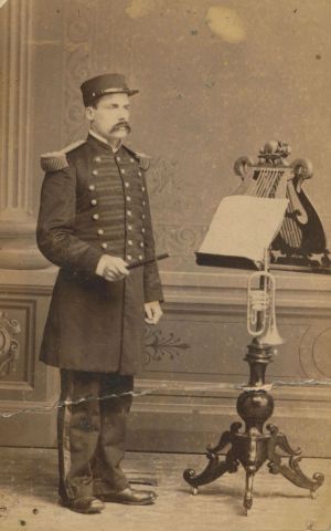 Photographie sépia d’un homme d’âge moyen. Il porte un uniforme et est debout devant un lutrin richement sculpté où reposent des partitions de musique et une trompette. Le nom et le titre du sujet sont écrits à la main au bas de la photo.
