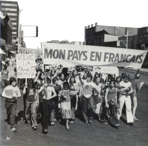 Photographie en noir et blanc d’un groupe d’adolescents manifestant en pleine rue. À l’avant, des jeunes portent une bannière qui se lit : «Mon pays en français» et une pancarte où est écrit : « Vive la francophonie ».
