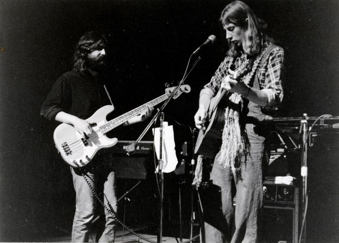 Black and white photograph of two young men, in long hair and casual clothes, on a stage, with amplifiers and an electric keyboard in the background. One of the musicans plays an electric guitar. The other plays an acoustic guitar and stands in front of a microphone.