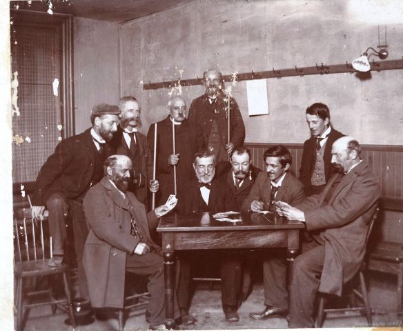 Photographie en noir et blanc d’un groupe d’hommes autour d’une table. Cinq hommes dont un religieux sont debout, tenant à la main, pour certains d’entre eux, une queue de billard et cinq hommes sont assis. Parmi ceux-ci, deux hommes ont des cartes dans les mains. La photographie est légèrement abîmée.