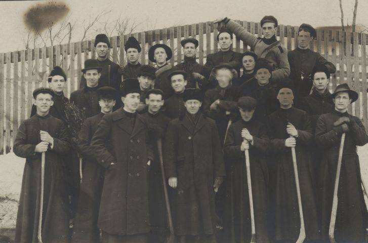 Photographie en noir et blanc, prise en hiver, d’un groupe d’une quinzaine de jeunes hommes, accompagnés de quelques adultes. Ils sont disposés en trois rangées, devant une clôture de bois. Les plus jeunes portent la soutane et tiennent des bâtons de hockey. Les adultes portent de longs manteaux d’hiver et des chapeaux de fourrure. L’un d’entre eux se tient devant le groupe, l’autre au centre de la photo, les deux autres sur les côtés.