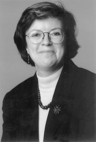 Photographie en noir et blanc, prise en studio, d’une jeune femme. Elle a les cheveux foncés, mi-longs, et elle porte des lunettes ainsi qu’un veston noir et un chandail blanc. Elle sourit à la caméra.