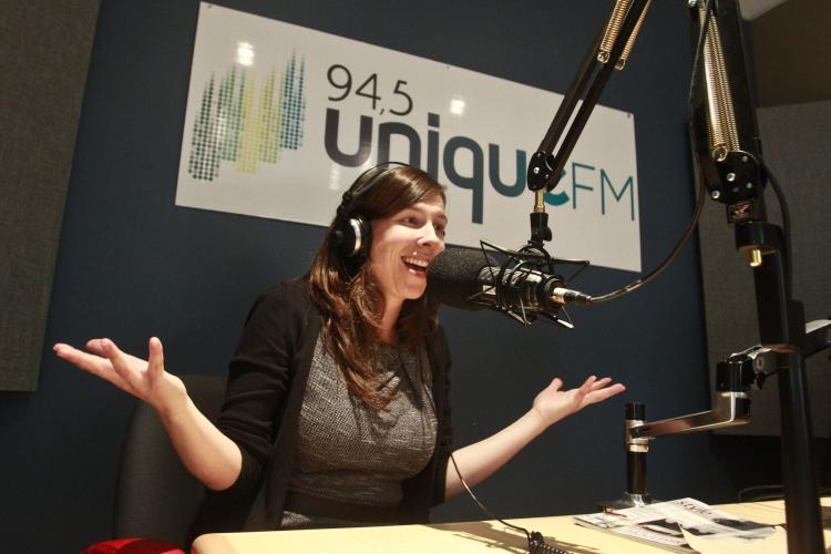 Photographie en couleur d’une femme souriante d’âge moyen, dans un studio de radio. Elle porte des écouteurs et parle devant un microphone suspendu. Elle est assise à une table, les bras levés et les mains ouvertes, les paumes tournées vers le haut. Derrière elle, une enseigne portant l’inscription : « 94,5 Unique FM ».