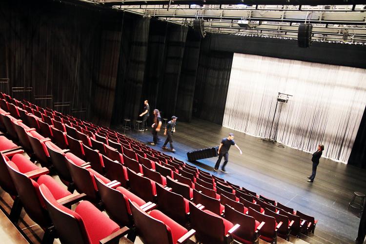 Photographie en couleur, prise de biais. Une salle de théâtre vue d’en haut, à partir des rangées de sièges rouge vif disposées en gradins. Cinq techniciens vêtus en noir travaillent sur la scène.