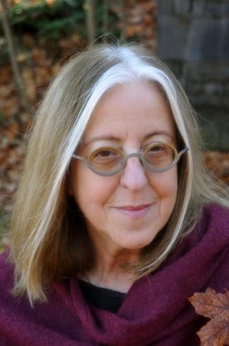 Photographie en couleur, prise à l’extérieur en automne, d’une femme d’âge mûr. Elle a les cheveux poivre et sel mi-longs. Elle porte des lunettes et une écharpe violette.  Elle sourit à la caméra.