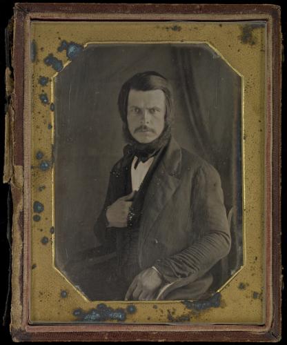 Photographie en noir et blanc, prise en studio, d’un homme à moustache au regard perçant, habillé d’un costume trois-pièces. Son bras gauche repose sur le bras du fauteuil où il est assis, et sa main droite est placée sous son gilet. La photographie est insérée dans un cadre doré, fortement abîmé.