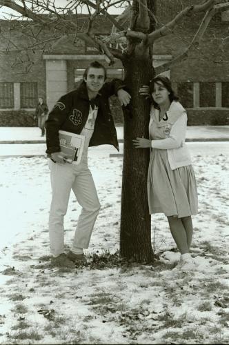 Photographie en noir et blanc d’un adolescent et d’une adolescente, dehors devant un édifice en brique. Ils s’appuient de façon décontractée contre un arbre en hiver. Il porte une veste en cuir et une chemise arborant une fleur de lys, tandis qu’elle porte une robe avec un grand col et une veste blanche.