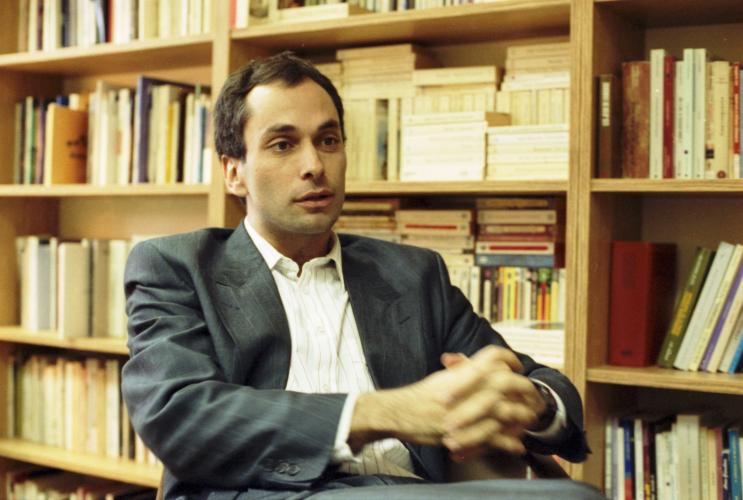 Photographie en couleur d’un homme d’âge moyen, vu de trois quarts. Il est assis, les mains jointes devant lui. Il a les cheveux bruns et la peau basanée. Il porte un costume sans cravate. À l’arrière-plan, une bibliothèque.