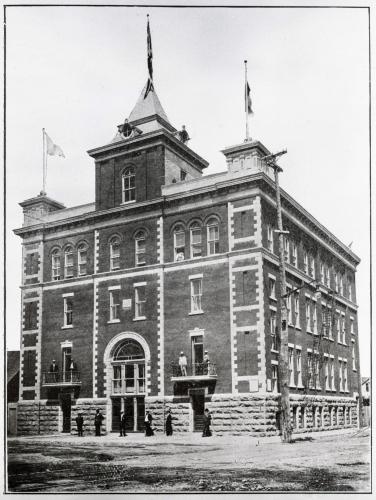 Photographie en noir et blanc d’un édifice en brique de quatre étages. La porte d’entrée est surmontée d’une tour, et deux tourelles sont placées de chaque côté à l’avant de l’édifice. Des drapeaux y flottent.  Des hommes circulent devant l’édifice et on peut voir quelques personnes sur les balcons du premier étage.