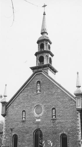 Photographie en noir et blanc d’une église en brique, surmontée d’un clocher et de deux tourelles. La façade de l’église compte trois portes, un œil-de-bœuf ainsi que trois statues.