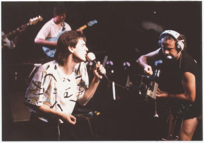 Colour photograph of a middle-aged man on stage, seen from the side. He sings with his eyes closed, holding a microphone in his left hand. In front of him, a cameraman; behind him, a guitarist.