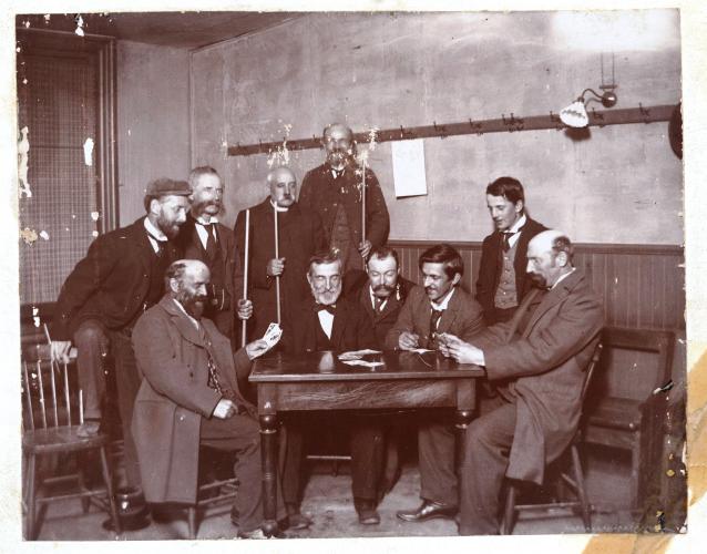 Photographie en noir et blanc d’un groupe d’hommes autour d’une table. Cinq hommes dont un religieux sont debout, tenant à la main, pour certains d’entre eux, une queue de billard et cinq hommes sont assis. Parmi ceux-ci, deux hommes ont des cartes dans les mains. La photographie est légèrement abîmée.